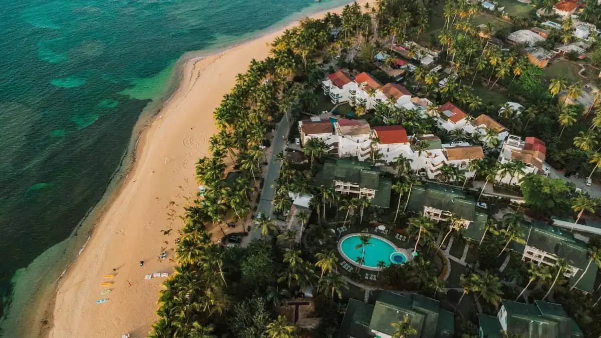 Vista aérea de una playa tropical con arena dorada, mar turquesa, palmeras y complejos turísticos con piscinas.