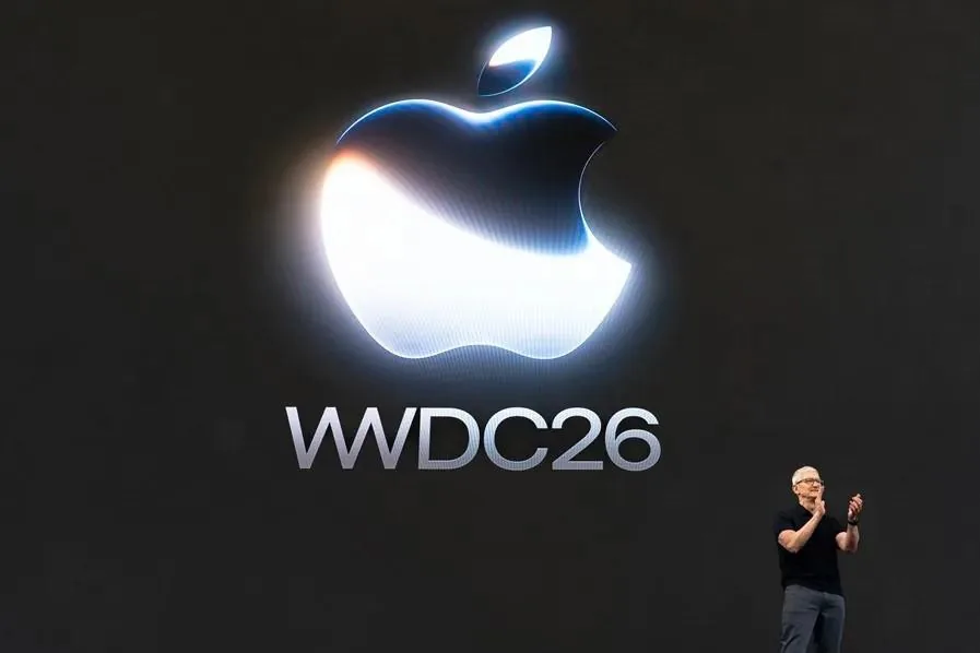 Presentador en el escenario frente a una gran pantalla oscura con el logotipo iluminado y el texto WWDC26.