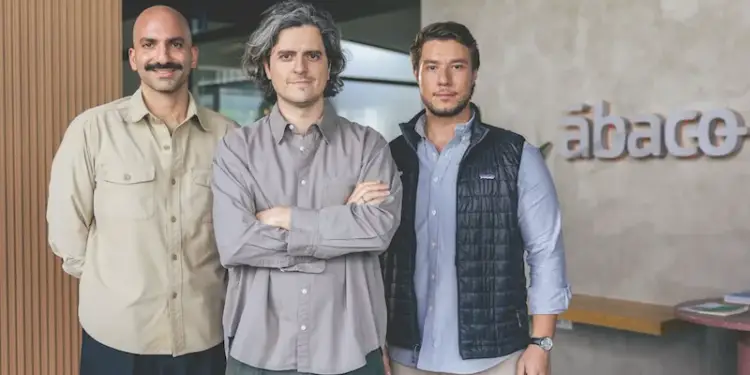 Tres hombres profesionales posan en una oficina moderna. Uno calvo con bigote, otro con cabello rizado y el tercero con chaleco. Fondo con logo 'ábaco'.