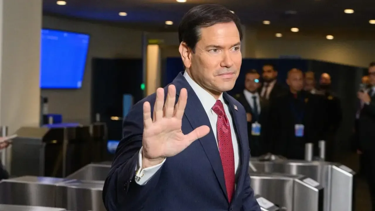 Hombre con traje azul y corbata roja levantando la mano en señal de saludo en un evento corporativo