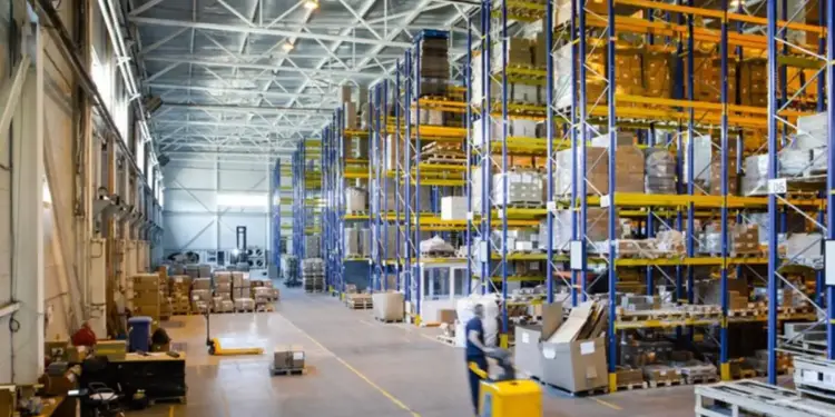 Interior de un gran almacén con estanterías altas llenas de cajas y palets. Un operario mueve mercancía con una transpaleta eléctrica.