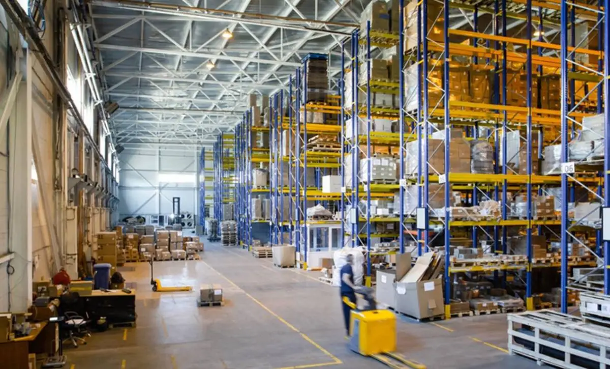 Interior de un gran almacén con estanterías altas llenas de cajas y palets. Un operario mueve mercancía con una transpaleta eléctrica.