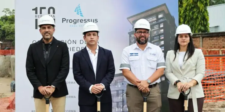 Cuatro profesionales con cascos y palas en ceremonia de primera piedra de un proyecto inmobiliario. Fondo con cartel de construcción.