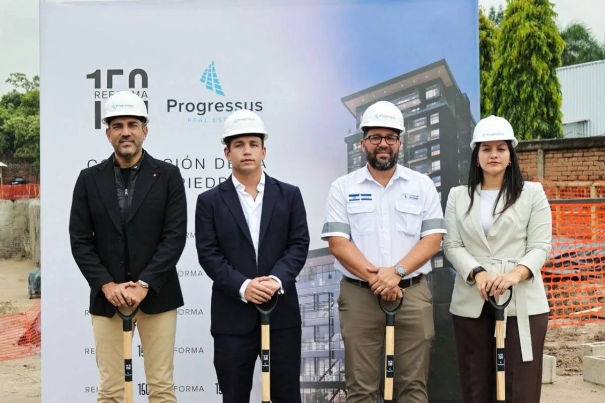 Cuatro profesionales con cascos y palas en ceremonia de primera piedra de un proyecto inmobiliario. Fondo con cartel de construcción.