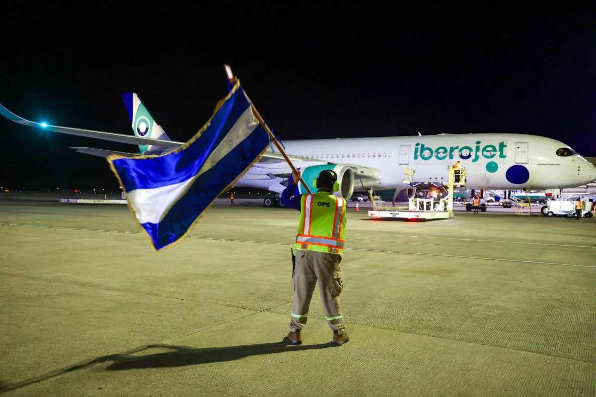Personal de tierra con bandera azul y blanca dirigiendo un avión Iberoject en pista de aeropuerto por la noche.