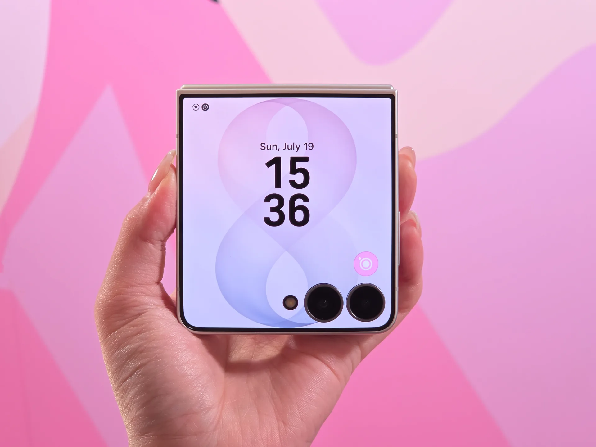 Mano sosteniendo un teléfono inteligente plegable compacto con pantalla exterior que muestra la hora sobre fondo rosa abstracto
