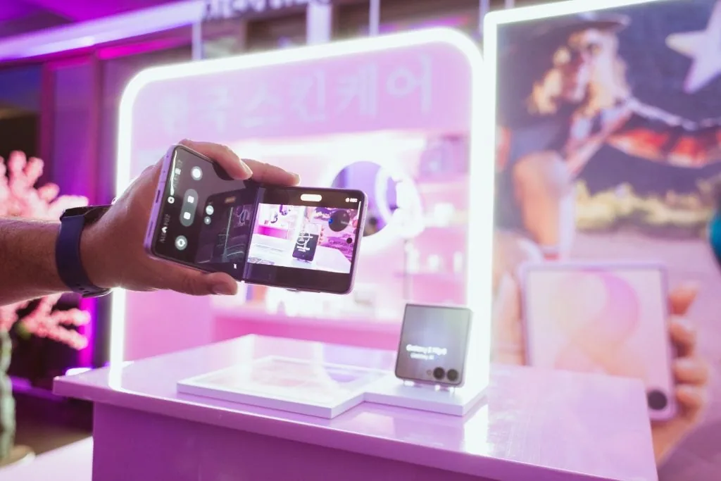 Mano de una persona sosteniendo un teléfono inteligente plegable en posición horizontal frente a un mostrador rosa iluminado.