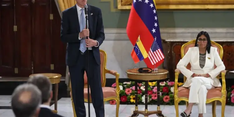Un hombre con traje azul hablando en un micrófono y una mujer sentada en una silla dorada junto a banderas de Venezuela y Estados Unidos.