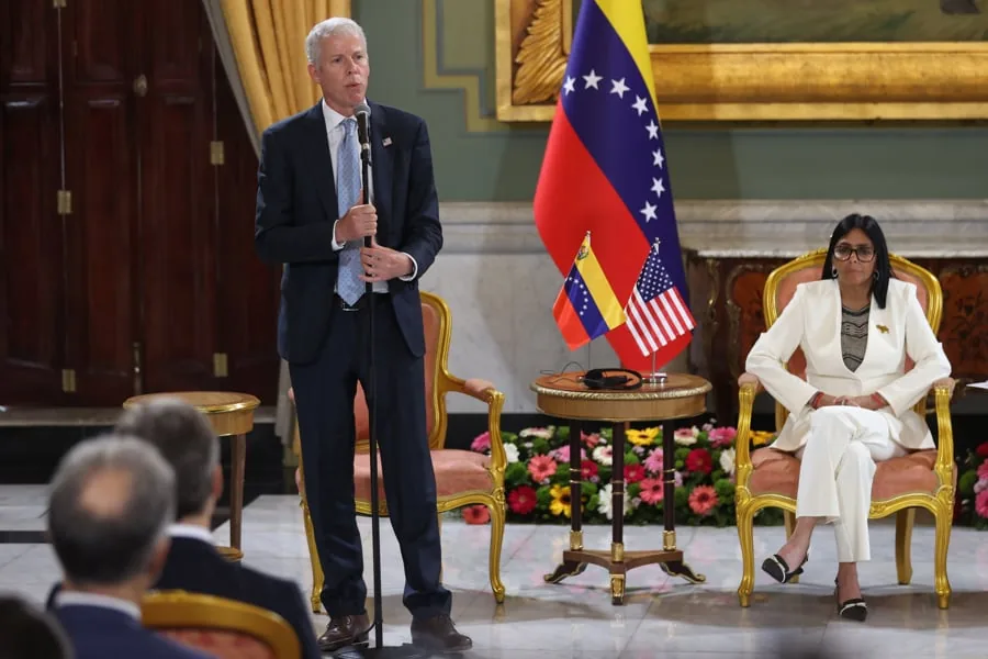 Un hombre con traje azul hablando en un micrófono y una mujer sentada en una silla dorada junto a banderas de Venezuela y Estados Unidos.