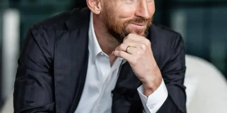 Hombre de negocios sonriente con traje oscuro, camisa blanca y barba, sentado en un sillón moderno.