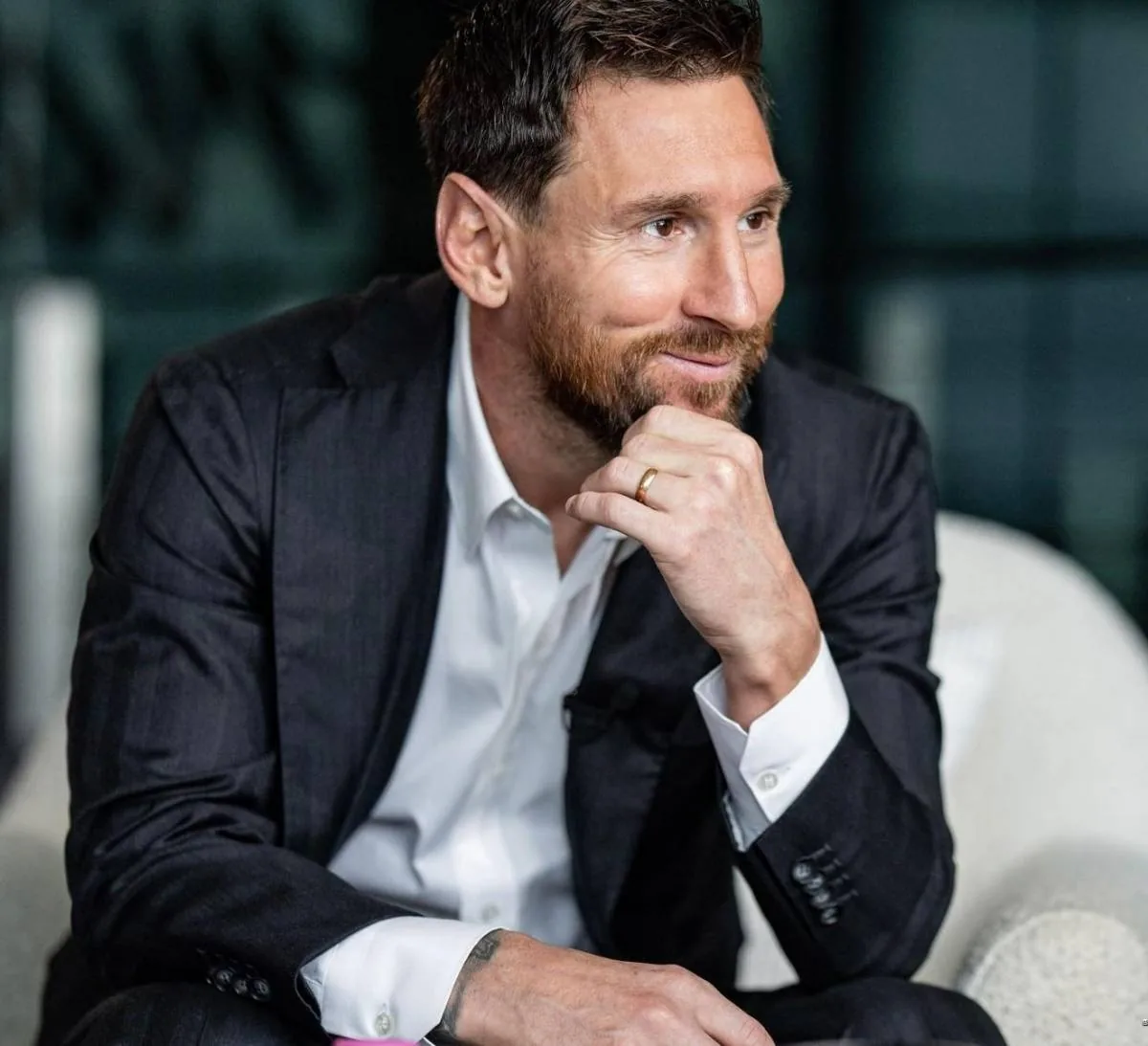 Hombre de negocios sonriente con traje oscuro, camisa blanca y barba, sentado en un sillón moderno.