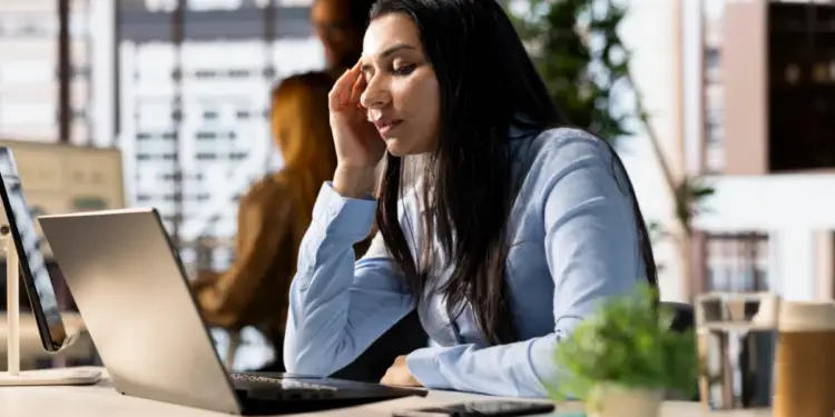 Mujer profesional con camisa azul claro, pensativa y con la mano en la sien, mirando su laptop en un escritorio de oficina. Hay un teléfono, planta y bebida.