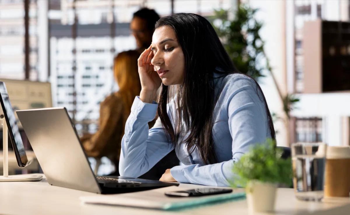 Mujer profesional con camisa azul claro, pensativa y con la mano en la sien, mirando su laptop en un escritorio de oficina. Hay un teléfono, planta y bebida.