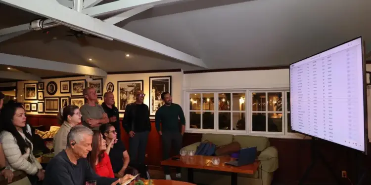 Un grupo de personas atentas mirando una pantalla grande con datos financieros en un espacio interior.