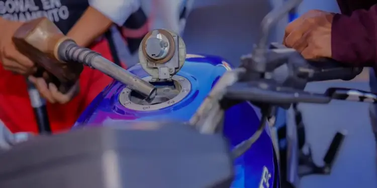 Primer plano de una persona repostando gasolina en el tanque de una motocicleta azul. Se observa la boquilla de la bomba y las manos del conductor.