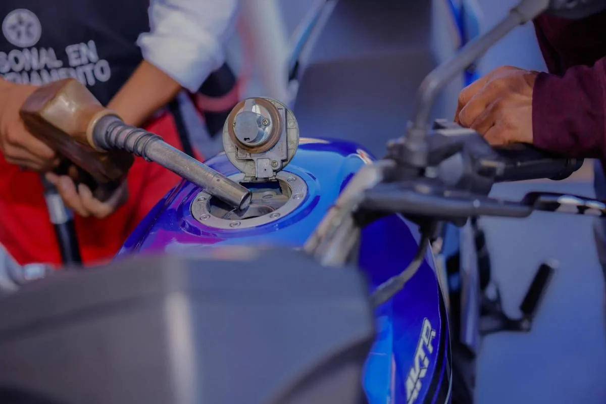 Primer plano de una persona repostando gasolina en el tanque de una motocicleta azul. Se observa la boquilla de la bomba y las manos del conductor.