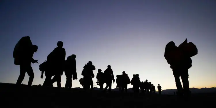 Siluetas de personas caminando en fila con mochilas y equipaje durante el atardecer.