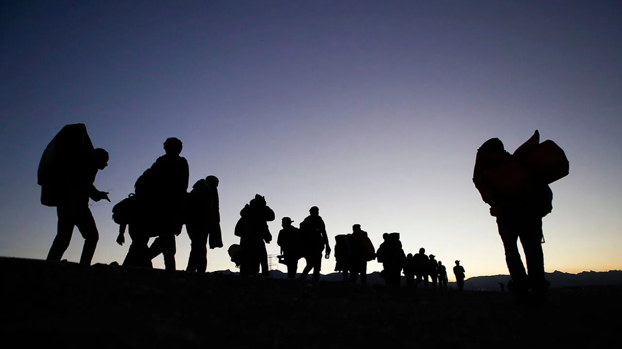 Siluetas de personas caminando en fila con mochilas y equipaje durante el atardecer.