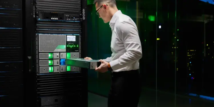 Profesional de TI gestionando un servidor en un centro de datos. Componentes electrónicos con luces LED verdes y marcas IBM y Schneider Electric visibles.