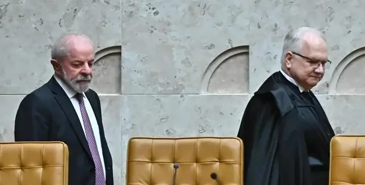 Dos hombres, uno con traje oscuro y corbata, otro con toga judicial, caminan en un entorno oficial con sillas de cuero.