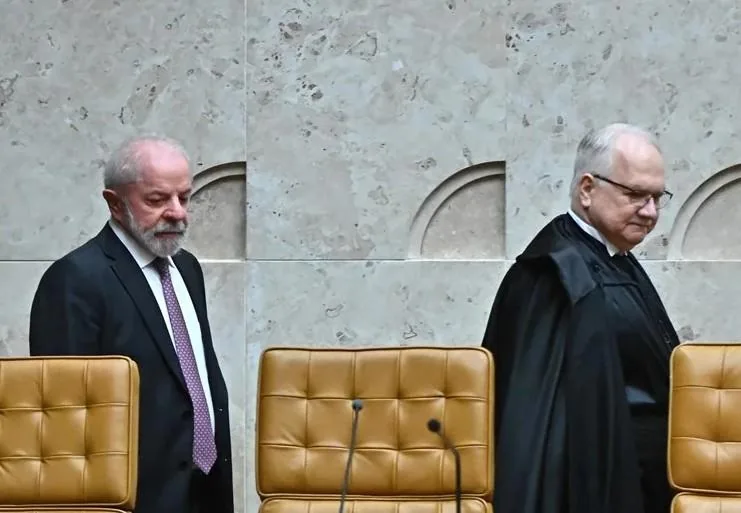 Dos hombres, uno con traje oscuro y corbata, otro con toga judicial, caminan en un entorno oficial con sillas de cuero.