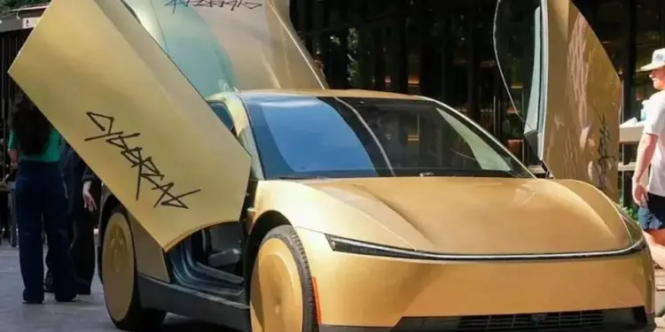 Coche futurista dorado con puertas abiertas hacia arriba, mostrando texto estilizado. Ruedas con cubiertas sólidas. Representa innovación automotriz.