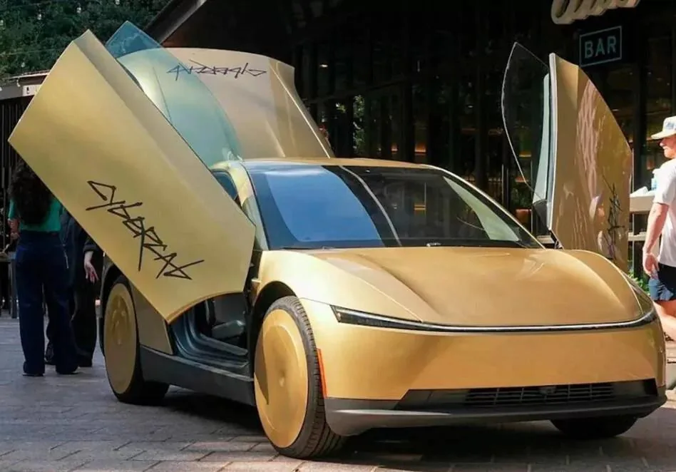 Coche futurista dorado con puertas abiertas hacia arriba, mostrando texto estilizado. Ruedas con cubiertas sólidas. Representa innovación automotriz.