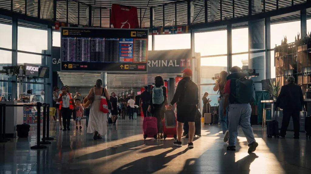 Personas caminando con equipaje en la amplia y luminosa terminal de un aeropuerto bajo pantallas de información de vuelos.