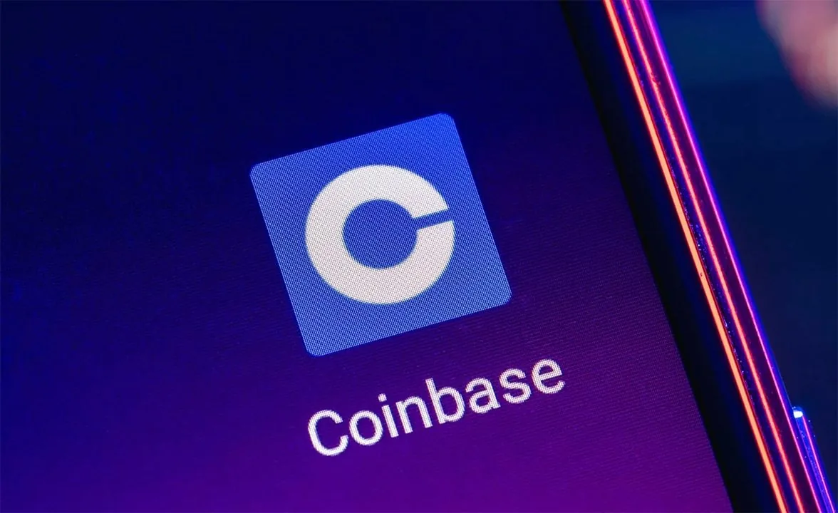 Primer plano del icono de la aplicación Coinbase con una 'C' blanca en un cuadrado azul, y el texto 'Coinbase' en una pantalla de smartphone. Fondo morado con reflejos de luz.