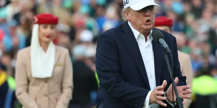 Hombre con traje azul y gorra blanca con las letras USA hablando ante un micrófono en un evento multitudinario.