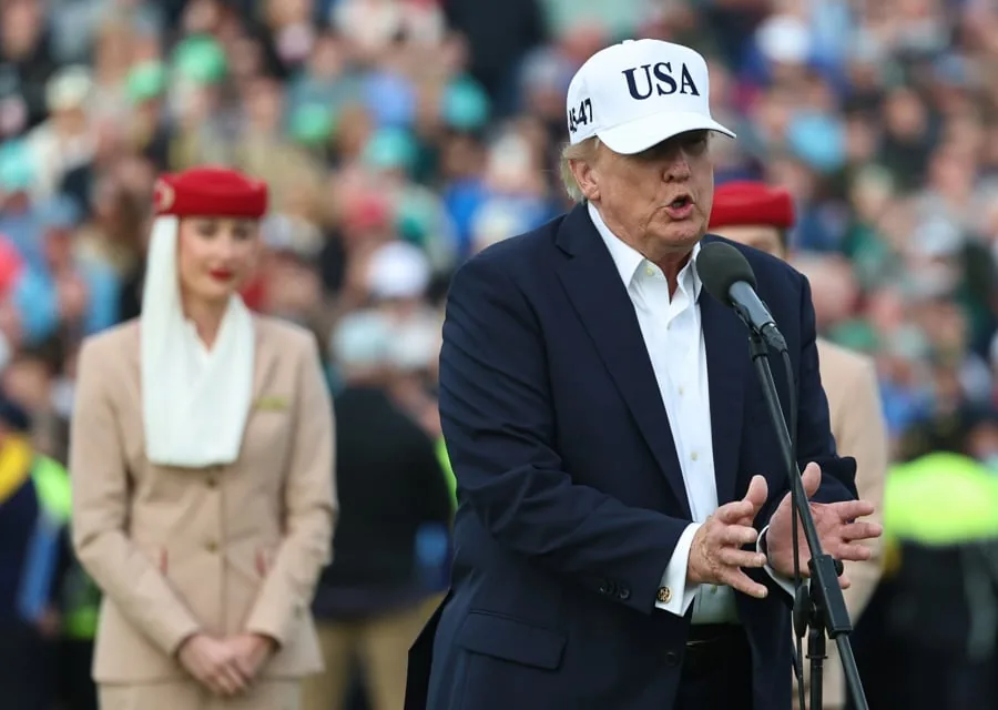 Hombre con traje azul y gorra blanca con las letras USA hablando ante un micrófono en un evento multitudinario.