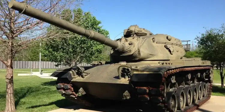 Tanque militar de color verde oliva con cañón largo, exhibido al aire libre sobre césped y concreto bajo un cielo azul claro.