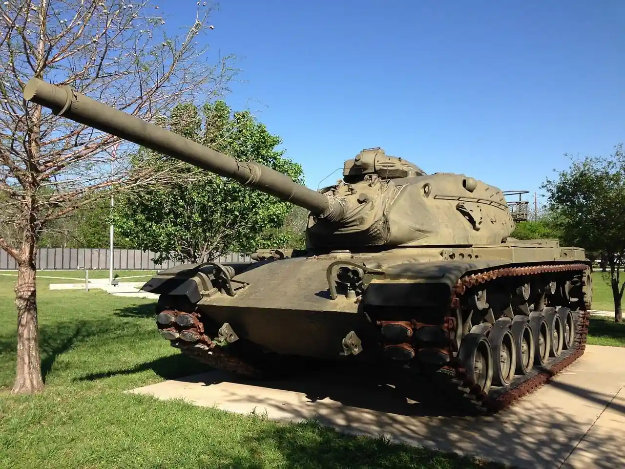 Tanque militar de color verde oliva con cañón largo, exhibido al aire libre sobre césped y concreto bajo un cielo azul claro.