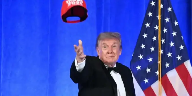 Donald Trump vistiendo traje formal frente a una bandera estadounidense y lanzando una gorra roja al aire.