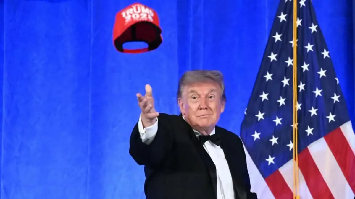 Donald Trump vistiendo traje formal frente a una bandera estadounidense y lanzando una gorra roja al aire.