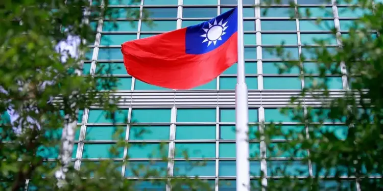 Bandera de Taiwán ondeando en un mástil frente a un edificio de oficinas con fachada de cristal y metal, con follaje verde en primer plano.
