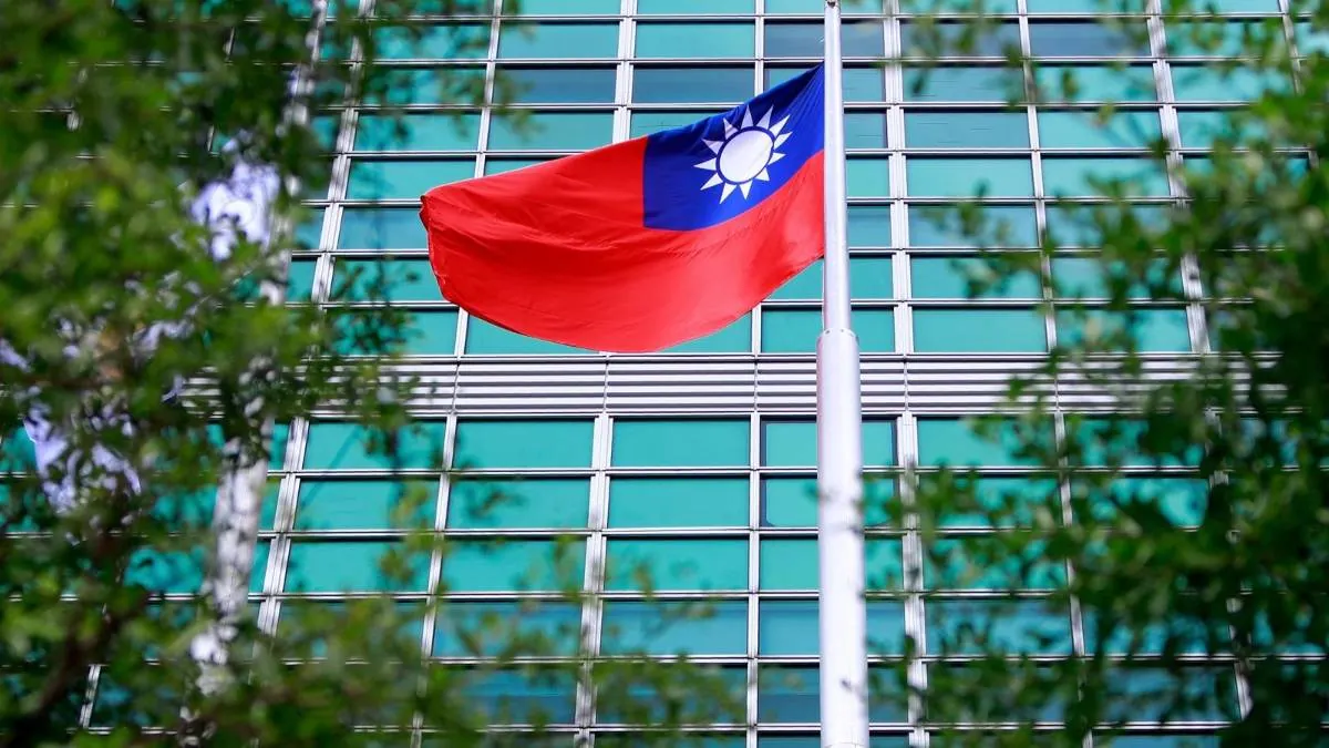 Bandera de Taiwán ondeando en un mástil frente a un edificio de oficinas con fachada de cristal y metal, con follaje verde en primer plano.