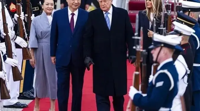 Dos líderes políticos caminando juntos sobre una alfombra roja acompañados por sus esposas y una guardia de honor con armas.