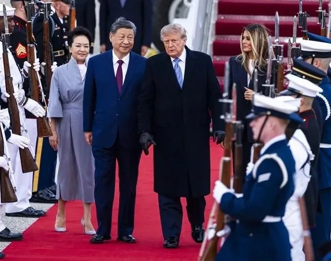 Dos líderes políticos caminando juntos sobre una alfombra roja acompañados por sus esposas y una guardia de honor con armas.
