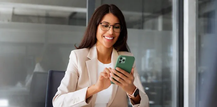 Mujer profesional con gafas y americana sonriendo mientras usa un smartphone en una oficina moderna.