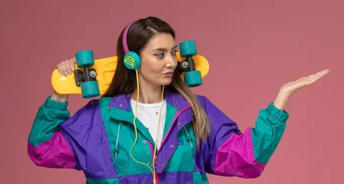 Mujer joven con chaqueta colorida (morado, turquesa, fucsia), auriculares rosas y turquesas, y patineta amarilla con ruedas turquesas al hombro. Fondo rosa.