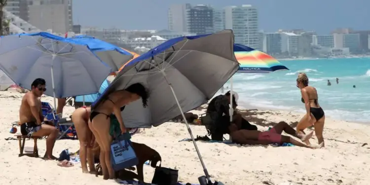 Personas relajándose bajo sombrillas en una playa de arena blanca con edificios al fondo.