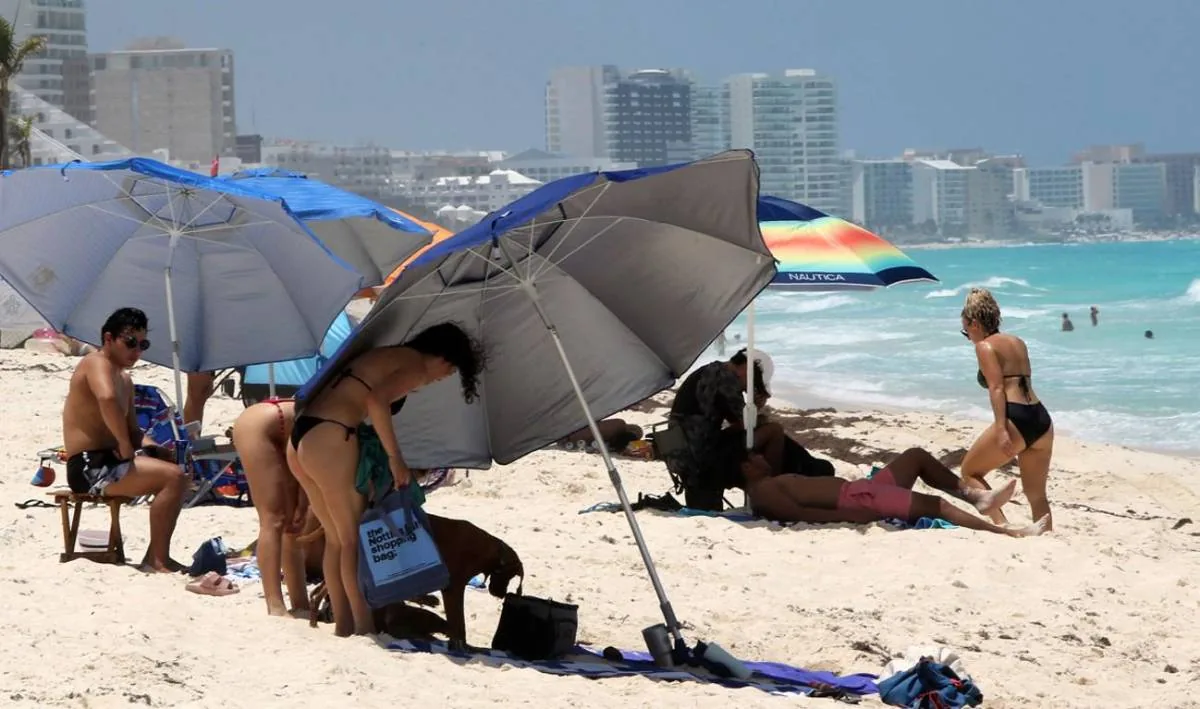 Personas relajándose bajo sombrillas en una playa de arena blanca con edificios al fondo.