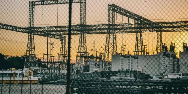 Vista de una subestación eléctrica y torres metálicas de alta tensión al atardecer detrás de una cerca metálica.