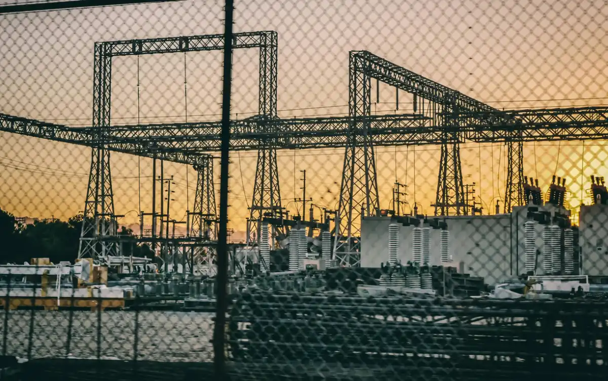 Vista de una subestación eléctrica y torres metálicas de alta tensión al atardecer detrás de una cerca metálica.