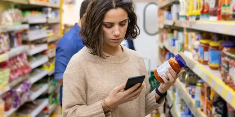 Mujer en supermercado comparando un frasco de salsa con su smartphone. Analiza información de producto y precios en el pasillo.