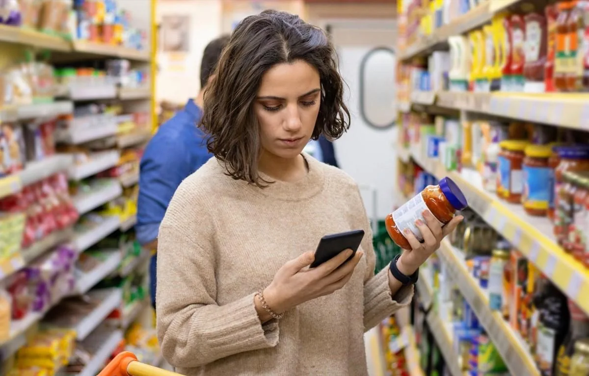 Mujer en supermercado comparando un frasco de salsa con su smartphone. Analiza información de producto y precios en el pasillo.