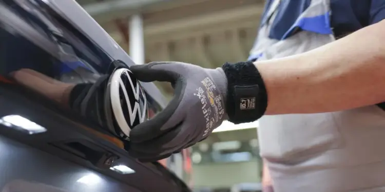 Mano enguantada de un operario de fábrica colocando el emblema de Volkswagen en la parte trasera de un vehículo oscuro, en una línea de ensamblaje.