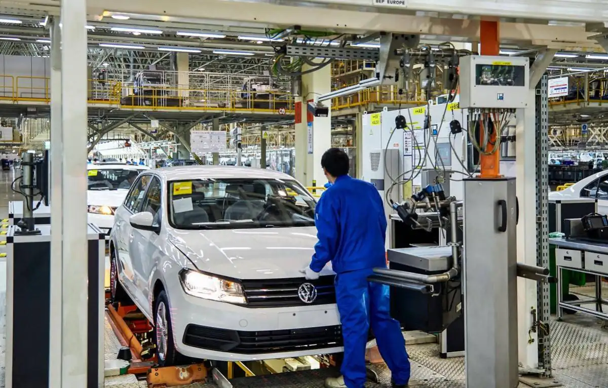 Operario con uniforme azul inspecciona un automóvil blanco en una línea de montaje e inspección industrial.