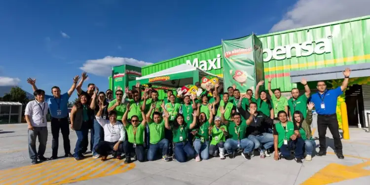 Gran equipo de empleados sonrientes en uniformes verdes posa frente a una tienda 'Maxi Pensa' con carteles de '¡Ya abrimos!' y 'Jocotenango', celebrando una apertura.